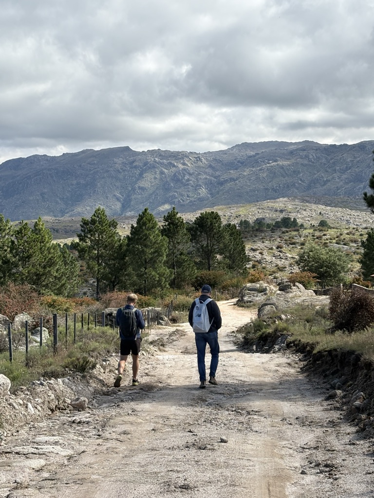 Two hikers are walking along a dirt path in a mountainous area. The landscape...