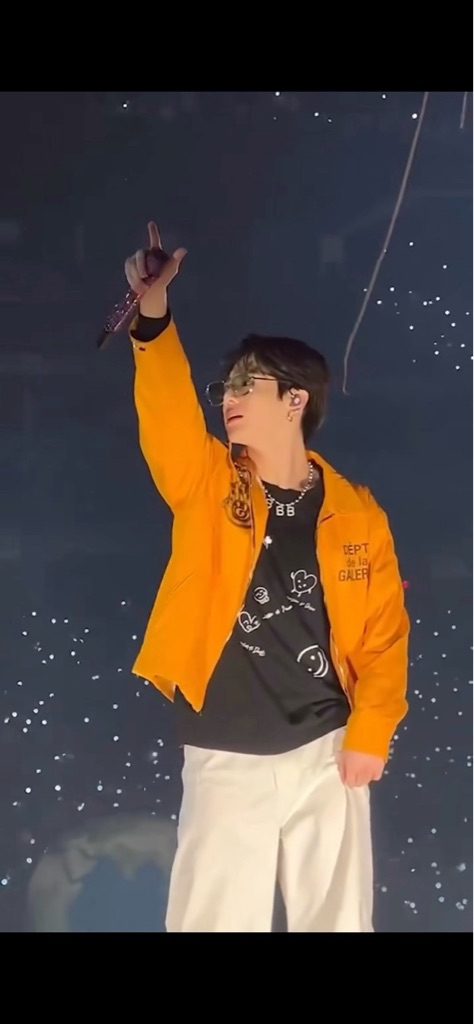 The image shows a performer on stage, wearing an orange jacket and a black sh...