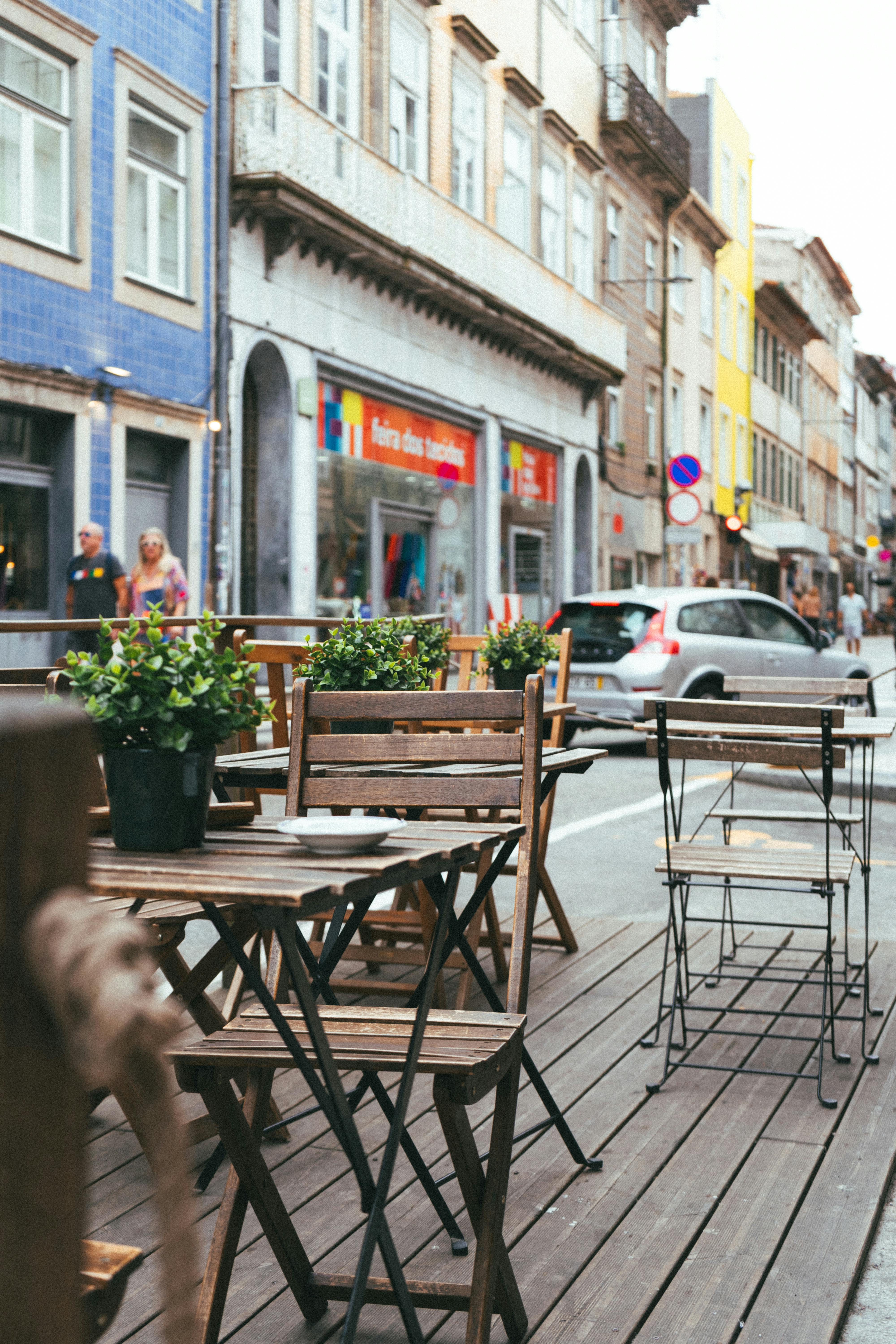 The image shows a street café with wooden tables and chairs. There are small...