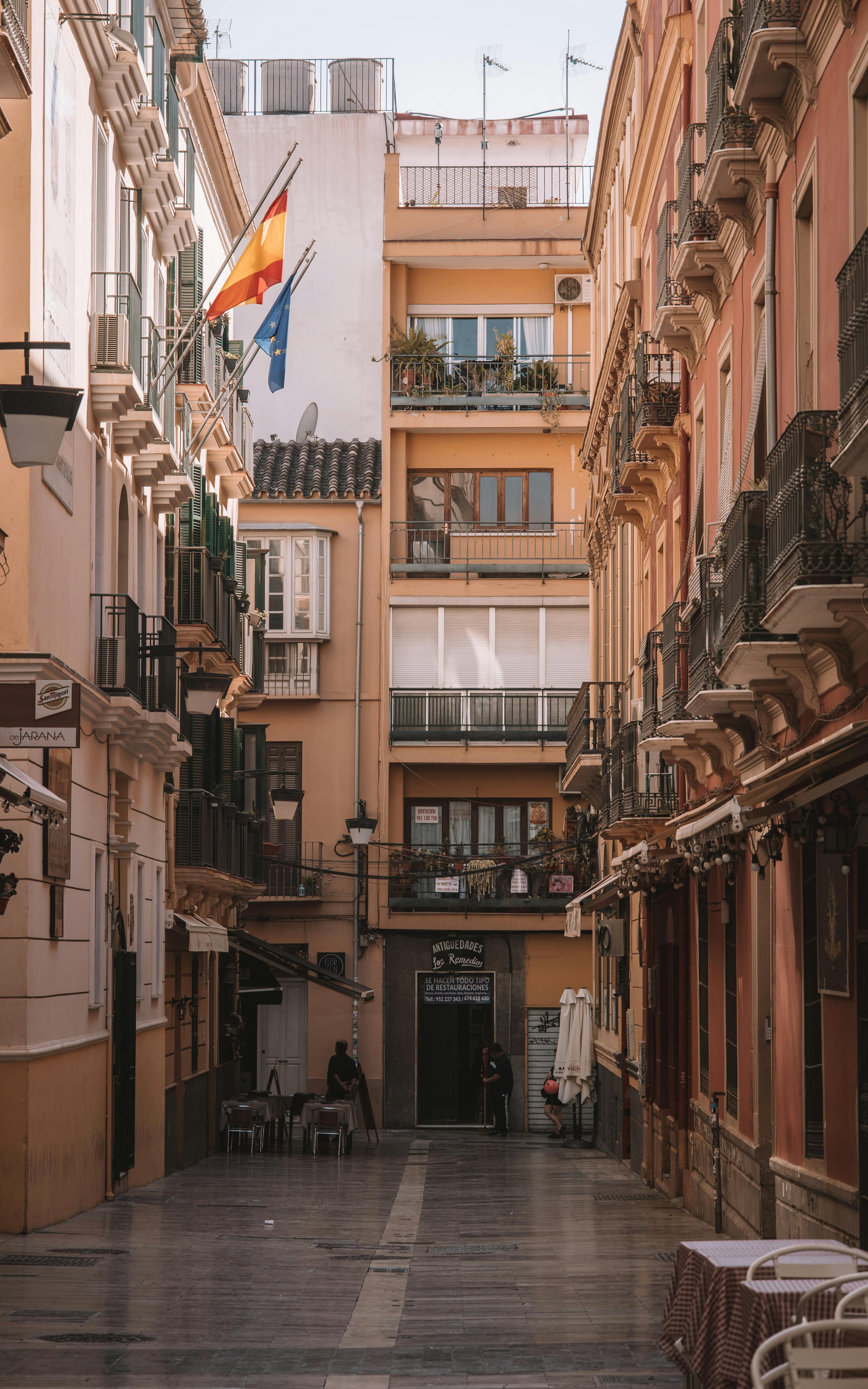 The photo shows a narrow street with colorful buildings. There are flags hang...